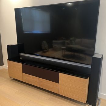 Full Spectrum Technology Group - Beautifully mounted Sony 98 Inch with receiver, surround sound, PS5, and apple TV in a Salamander floating cabinet - A large flat-screen TV is mounted on a modern wooden TV stand with a combination of light and dark wood finishes. Two tall speakers are positioned on either side of the TV, and the stand features multiple compartments for storage. The room has light-colored wooden flooring and neutral wall colors, with a reflection of a sofa and coffee table visible on the TV screen. - Home Theatre Installation Near Me - Concord, California
