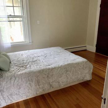 Hope Woodworks - You can see the single door opening to the old closet where he then knocked the wall down to create the reach-in closet. - The image shows a bedroom with a bed covered in a white patterned bedspread, positioned near a window with sheer curtains. The room has beige walls, a baseboard heater along the wall, and a dark wooden door. The hardwood floor is visible, and the room appears to be well-lit with natural light from the window. - General Contractors Near Me - Woonsocket, Rhode Island