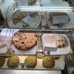 Cookies and cakes - The bakery case contains multiple baked goods arranged on trays with paper liners. Items include blueberry crumb cake, sour cream crumb cake, almond croissants, cranberry scones, and cookies. Each item has a label with its name and price, such as $3.25 for almond croissants and $4.00 for blueberry crumb cake. The display is behind a glass front, with some reflection visible on the surface.