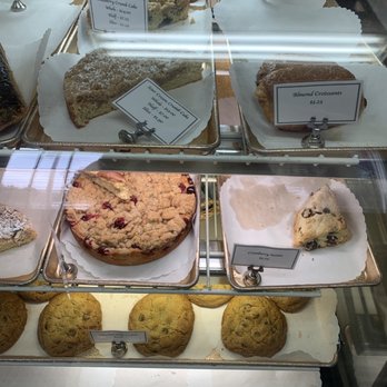 A-Frame Bakery - Cookies and cakes - The bakery case contains multiple baked goods arranged on trays with paper liners. Items include blueberry crumb cake, sour cream crumb cake, almond croissants, cranberry scones, and cookies. Each item has a label with its name and price, such as $3.25 for almond croissants and $4.00 for blueberry crumb cake. The display is behind a glass front, with some reflection visible on the surface. - Bakeries Near Me - Williamstown, Massachusetts