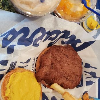Culver's - Yummy - burger, fries, rootbeer float and a free yogurt with peaches. - The image shows a fast food meal on a blue tray, including a burger with yellow mustard, a fried beef patty, and a side of French fries. Two beverages are also present: one appears to be a milkshake or smoothie with a straw, and the other is a dessert or fruit topping with a spoon. The tray is lined with branded paper, and the food items are arranged for easy access. - Burgers Near Me - St. Cloud, Florida