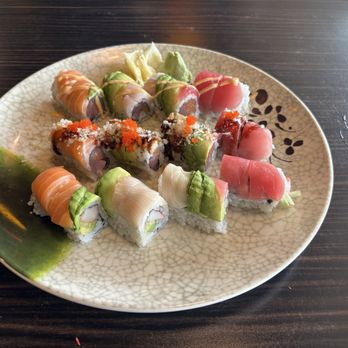 Osaka - Rainbow, Tiger and Double 007 rolls. - A round ceramic plate contains an assortment of sushi rolls, including varieties topped with slices of fish, avocado, and sauces. Some rolls are garnished with fish roe and drizzled sauces, with shredded lettuce or cabbage on the side. The plate is placed on a dark wooden table, and the sushi presentation features colorful ingredients and decorative toppings. - Asian Fusion Near Me - Bloomfield, Connecticut