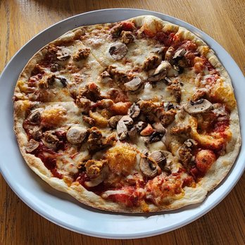 MOD Pizza - The Jasper - A round pizza topped with sliced mushrooms, crumbled sausage, and melted cheese is served on a white plate. The pizza has a thin crust and is set on a wooden table, with another pizza partially visible in the background. - Fast Food Near Me - Dunkirk, Maryland