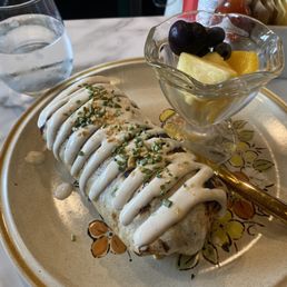 Lil Donkey - A burrito with white sauce and chopped herbs is placed on a decorative ceramic plate with a floral pattern. Next to it, a small glass bowl contains pineapple, grapes, and orange segments. A glass of water is positioned behind the plate on a marble countertop. The setting appears to be a casual dining environment, possibly a restaurant or cafe.