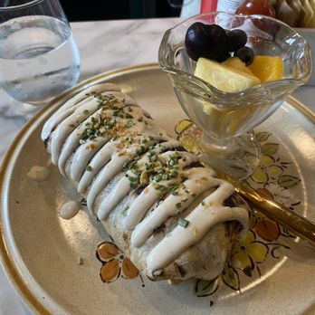 Toad in the Hole - Lil Donkey - A burrito with white sauce and chopped herbs is placed on a decorative ceramic plate with a floral pattern. Next to it, a small glass bowl contains pineapple, grapes, and orange segments. A glass of water is positioned behind the plate on a marble countertop. The setting appears to be a casual dining environment, possibly a restaurant or cafe. - Breakfast & Brunch Near Me - Eagle River, Wisconsin