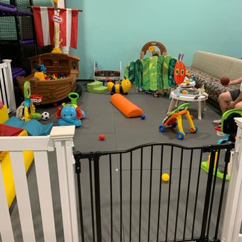 Wisco Kidz Place - An indoor children's play space features a variety of toys, including a ball pit, a small fence, a tunnel, and soft play equipment. The area has a light blue wall, a couch with a child sitting on it, and a padded floor for safety. Toys such as a bee-shaped ride-on, a soccer ball, and a colorful caterpillar-shaped play structure are visible, creating a safe and engaging environment for children. - Playgrounds Near Me - Madison, Wisconsin