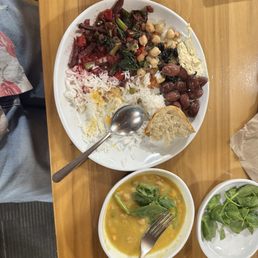 Idk - The image shows a wooden table with a large white plate holding rice, chickpeas, olives, salad, and a piece of bread. A metal spoon rests on the rice. Next to the plate is a bowl of yellow soup with leafy greens and a fork. A small bowl of fresh greens is also visible. The setting appears to be a casual dining environment with a notebook in the background.
