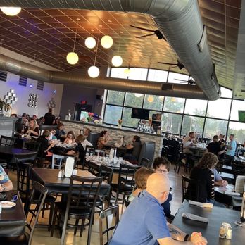 Serventi's - Interior photo - The image shows the interior of a restaurant with numerous diners seated at tables, some engaged in conversation and others eating. The space features large windows allowing natural light, modern ceiling lighting with round pendant fixtures, and visible ductwork on the ceiling. The decor includes a stone accent wall and contemporary wall art. The restaurant appears busy, with a mix of groups and individuals dining. - Italian Near Me - Butler, Pennsylvania