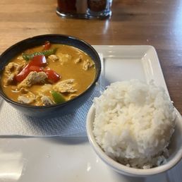CU1. Panang Curry Dinner - A bowl of Thai curry with red and green peppers and chicken, accompanied by a bowl of white rice, on a white tray at a restaurant.