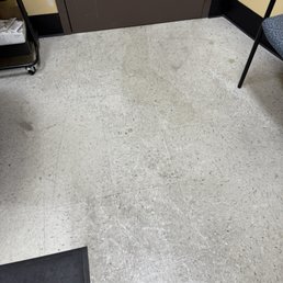 The image depicts a section of an indoor floor with light-colored tiles that have visible dirt, scuff marks, and some debris. A brown door is positioned at the top center, with a black baseboard running along the wall. To the right, part of a chair with a patterned cushion is visible, and on the left, a small cart with items, possibly papers or packaging, is partially seen. The setting appears to be a utilitarian or office space.