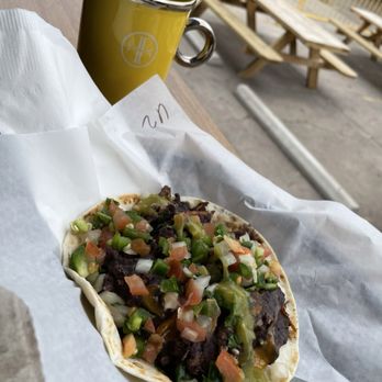 Lazarus Brewing - Bean, Frijoles, Egg and Cheese - A taco filled with beef, chopped vegetables, and salsa is placed on a paper-lined tray outdoors, with a yellow mug and picnic tables in the background. - Breweries Near Me - Austin, Texas