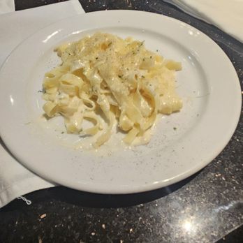 BrickTop's - Side dish of fettuccine - A white round plate contains cooked fettuccine pasta coated in a creamy white sauce, garnished with grated cheese and black pepper. The plate is set on a dark, speckled restaurant table with a glass of water, salt and pepper shakers, and a napkin with a utensil. The background shows part of the restaurant setting with additional condiments and tableware. - New American Near Me - Frontenac, Missouri