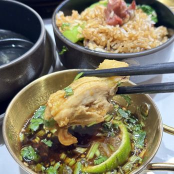 Coco Hotpot - A piece of cooked chicken is held with chopsticks above a small bowl containing soy sauce, chopped herbs, and lime slices. In the background, there is a bowl of rice with vegetables and a dark cup, indicating a meal setting. The image captures a close-up of the dipping process, highlighting the ingredients and presentation. - Hot Pot Near Me - Flushing, New York