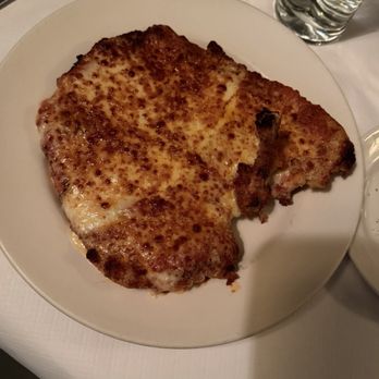 Hendrick's Tavern - A photograph shows a large, breaded chicken cutlet that has been baked or fried, placed on a white ceramic plate. The chicken has a golden-brown, crispy coating and appears to be a main course dish. The setting includes a glass of water, a fork, and a knife on a white tablecloth, indicating a restaurant dining environment. - Bars Near Me - Roslyn, New York