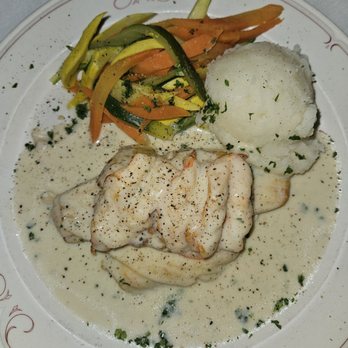 Casa Bella - A plate contains a fish fillet covered in a white creamy sauce, accompanied by a serving of mashed potatoes and a side of sautéed mixed vegetables including carrots, zucchini, and yellow squash. The dish is garnished with chopped herbs and black pepper, presented on a white plate with a decorative red border. - Italian Near Me - Scranton, Pennsylvania