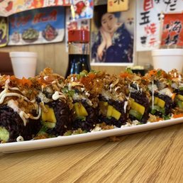 French Kiss Roll - A rectangular white plate contains several sushi rolls with dark rice, topped with drizzles of mayonnaise and other sauces, chopped green onions, and crispy toppings. The sushi is arranged in a line on a wooden table, with cups and a soy sauce bottle visible in the background, along with Japanese posters and decorations on the wall.