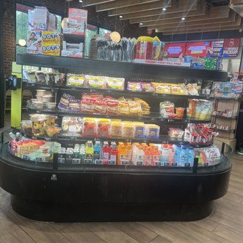 Sheetz - Breakfast items - A black refrigerated display case with multiple shelves holds a variety of snacks, drinks, and breakfast items inside a convenience store. The top shelf contains packaged snacks and breakfast foods, while the lower shelves are stocked with bottled beverages, including juices and water. The store has a modern interior with wooden ceiling accents, brick walls, and large windows allowing natural light. The background shows additional store shelves and signage. - Convenience Stores Near Me - Swiftwater, Pennsylvania