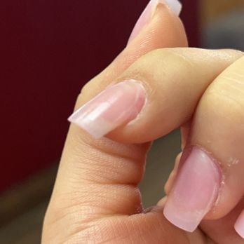 Julia's Nails - UNEVEN APPLICATION AND ALLREADY CHIPPING - The image shows a close-up of a person's hand with nails featuring a French manicure. The nails have a clear or pale pink base with white tips, but the application appears uneven, and some nails show signs of chipping. The background is blurred, focusing on the nails and hand, which are positioned to highlight the imperfections in the manicure. - Nail Salons Near Me - Phoenix, Arizona