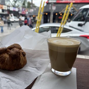 Augusta Coffee - Coffee and croissant cookie - On a wooden table outside a coffee shop, there is a croissant wrapped in white paper, a glass of water, and a latte with foam art in a clear glass. The background shows a busy street with pedestrians, parked cars, and a white van, with storefronts and people walking along the sidewalk. - Coffee Roasteries Near Me - Toronto, Ontario
