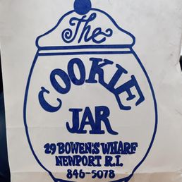 a sign for the cookie jar - The image shows a white paper bag with a blue printed logo and text. The logo features a stylized jar with a lid and a small round handle on top. Inside the jar outline, the words "The Cookie Jar" are written, with "The" in cursive and "Cookie Jar" in bold, uppercase letters. Below the logo, the address "29 Bowens Wharf, Newport, RI" and the phone number "846-5078" are printed. The bag appears to be crumpled and is likely used for carrying baked goods from The Cookie Jar in Newport, RI.
