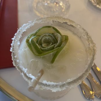 Odelia - Coconut margarita - A beverage in a glass with a salted rim is topped with a cucumber rose garnish. The drink is placed on a white plate, with a straw visible. In the background, there are wine glasses and silverware, indicating a dining setting. The presentation suggests a crafted cocktail, possibly at a restaurant or bar. - Coffee & Tea Near Me - Mandeville, Louisiana