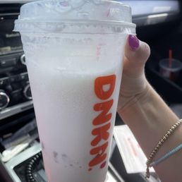 I ordered a fucjing iced chai - A person with purple nail polish and bracelets holds a large Dunkin' iced beverage with a straw, inside a car. The drink appears to be a milkshake or iced coffee, with a clear plastic lid and a straw. The background shows the car's dashboard and interior, including the radio and air vents.