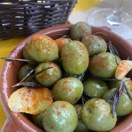 Olives - A terracotta bowl contains green olives sprinkled with red paprika, garnished with lemon slices, on a yellow table with a wine glass and basket in the background.