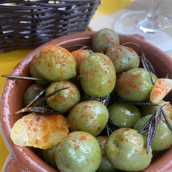 Maite - Olives - A terracotta bowl contains green olives sprinkled with red paprika, garnished with lemon slices, on a yellow table with a wine glass and basket in the background. - Colombian Near Me - Brooklyn, New York