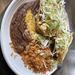 Beef Taco - A large white plate holds a Mexican meal including a shredded beef taco with lettuce and cheese, a bean tostada topped with shredded lettuce and cheese, Mexican-style rice, and refried beans. The food is arranged on a wooden table, with a spoon partially visible at the edge of the plate.
