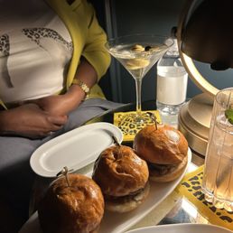 Sliders - Three sliders with shiny buns and fillings are arranged on a white oval plate on a table. The table also holds a martini glass with an olive garnish, a bottle of water, and a glass with a beverage and mint leaves. A person wearing a yellow jacket and a watch is seated nearby, with part of their torso visible. The setting appears to be a restaurant or bar environment.