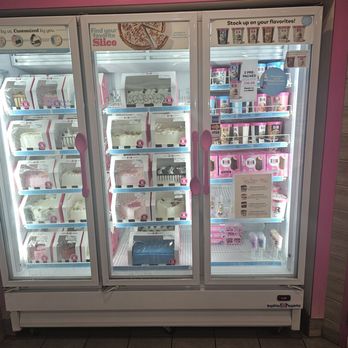 Baskin-Robbins - Freezer with ice cream cakes and ready made pints of ice cream - A large glass display freezer with three sections showcases ice cream cakes and pints at Baskin-Robbins. The left and middle sections contain boxed ice cream cakes on shelves, while the right section displays pints of ice cream with promotional signage. The freezer has pink handles and is situated against a wall with pink and wood paneling accents. Signage on the top highlights flavor options and promotional offers. - Desserts Near Me - Modesto, California