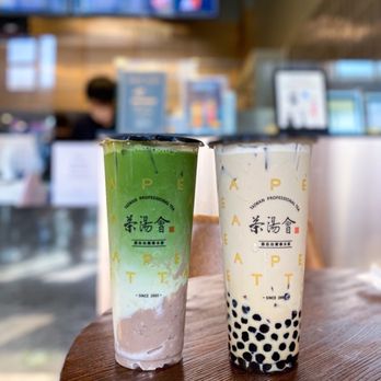 TP TEA - Taro Matcha latte & Tie Guan Yin Milk Tea with Boba and QQ Noodles - Two bubble tea drinks are positioned on a wooden table inside a cafe. The drink on the left has a green tea color with a green top, while the drink on the right is a creamy white with black tapioca pearls at the bottom. The cups feature branding and text, and the background shows a blurred cafe interior with a person, menu screens, and digital ordering devices. - Coffee & Tea Near Me - Berkeley, California