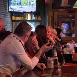 A group of people sit at a bar counter in a restaurant or pub, with drinks and snacks, watching a TV screen showing a sports game.