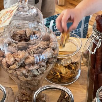Squeaky Bea’s - Cute treats in cute containers - A large glass jar containing dried ginger roots is on a wooden table, with a person reaching into a bowl of dried fruit nearby. - Pet Stores Near Me - Chicago, Illinois