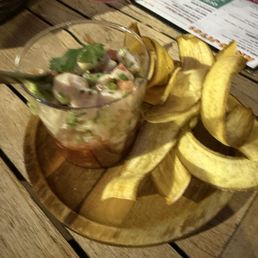 Ceviche - A glass bowl of ceviche garnished with herbs is placed on a wooden tray alongside plantain chips, on a rustic wooden table.