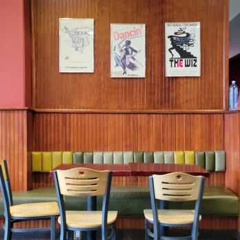 Project Burger - The image shows a dining area with a wooden-paneled wall decorated with three posters, including one with a dancer and another with a stylized figure. A cushioned bench with green and yellow sections runs along the wall, paired with a red table. In front of the bench, there are three wooden chairs with beige seats and black frames. The floor is dark wood, and a water bottle is visible on the right side of the image. The setting appears to be a casual dining space or cafe. - American Near Me - Alameda, California