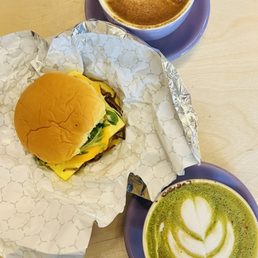 Breakfast Sandwich - A breakfast sandwich with cheese, greens, and bacon on a bun, accompanied by a cappuccino with foam art and a matcha latte with a tulip design.