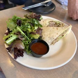 Avocado wrap - A white plate holds a cut wrap filled with meat and vegetables, accompanied by a side of mixed greens and a small black cup of sauce. The plate is on a wooden table in a restaurant setting, with utensils, a glass of water, and a napkin visible nearby.