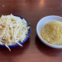 Bean Sprout Kimchi - The image shows a small blue bowl filled with bean sprout kimchi topped with sesame seeds, alongside a white dish containing a scoop of sweet corn cake. Both dishes are set on a wooden table, suggesting a casual dining setting at a restaurant that offers Korean and fusion cuisine.