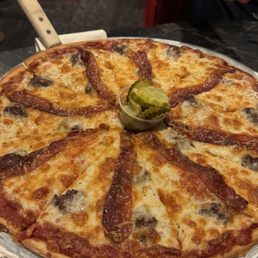 Cheeseburger - A pizza topped with bacon, cheese, and mushrooms, served on a metal tray with a small container of pickles in the center.