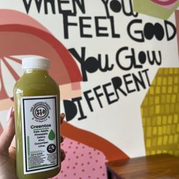 A person is holding a bottle of green juice labeled 'Greentox' with ingredients including kale, apple, pineapple, celery, kiwi, cucumber, and lime, in front of a colorful wall. The wall features a motivational quote that reads, 'When you feel good, you glow different,' with bold black letters and abstract shapes in pink, orange, yellow, and green.