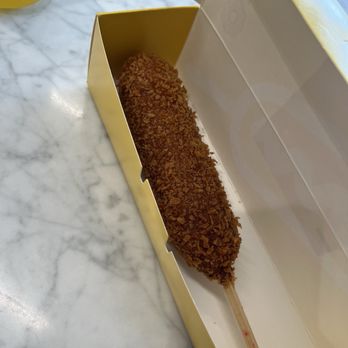 Mochinut - A deep-fried snack on a wooden stick, coated with a crunchy, golden-brown breadcrumb layer, is positioned inside a yellow and white box. The box is open, revealing the snack resting on a marble surface. The background includes a yellow object and a marble tabletop, indicating a casual dining or snack setting. - Hot Dogs Near Me - Columbus, Ohio