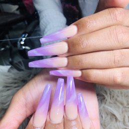 A set of long, almond-shaped acrylic nails features a purple and nude gradient design, held by two hands with well-manicured skin.