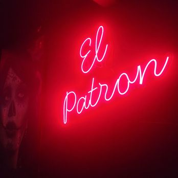 El Patron - A neon sign with pink cursive lettering spells out "el patron" on a dark wall, illuminated against the background. To the left, a partial view of a colorful face painting or mural is visible, featuring a face with detailed designs. The setting appears to be indoors, possibly in a bar or restaurant with decorative lighting. - Mexican Near Me - Flushing, New York