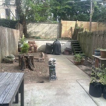 Native NY Gardens - Before - A backyard features a concrete patio area with potted plants along the right fence, a tree stump in the center, and a barbecue grill at the back. There are garden beds with bricks and a small staircase leading to a higher level. The space is enclosed by wooden fences on both sides, with trees and buildings visible beyond. - Landscaping Near Me