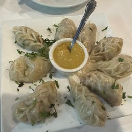 Veggie Vegetable Steamed MoMo - The image shows a square white plate with ten steamed vegetable momos arranged around a small bowl of yellow dipping sauce. The momos are garnished with chopped herbs, and a spoon is placed in the sauce. The setting suggests a restaurant serving traditional Nepalese cuisine, specifically vegetarian momos with a dipping sauce.