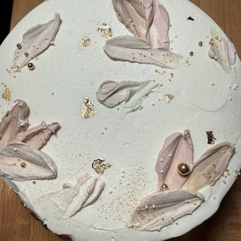 The Evercake - A round cake features a smooth white surface with decorative dollops of pastel-colored meringue-like frosting in shades of pink, beige, and light green. Gold leaf accents and small edible beads are scattered across the top, creating an elegant appearance. The cake is placed on a wooden surface, and the decorations are arranged in a seemingly random pattern, emphasizing a sophisticated and artistic design. - Custom Cakes Near Me