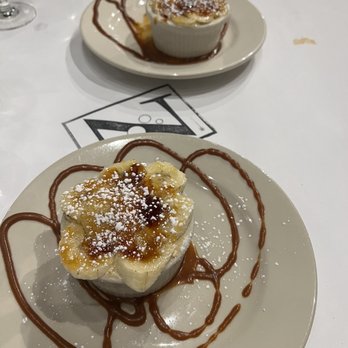 Zarletti - Banana Semi - freddo - Banana foster dessert presented in a ramekin with caramel sauce, powdered sugar, and chocolate drizzle on a white plate at a restaurant. - Italian Near Me - Milwaukee, Wisconsin