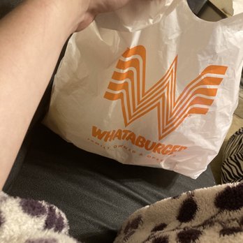 Whataburger - nothing - A person holds a white Whataburger paper bag with orange logo and text, sitting on a dark surface, with part of their clothing visible. - Burgers Near Me - Tomball, Texas