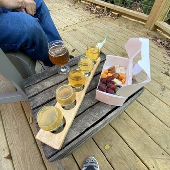 Three Falls Cider - Beer $6, flight $10, charcuterie box $13 - The image shows a cider tasting arrangement on a wooden deck, featuring five glasses of cider placed on a wooden tasting board. A person wearing jeans and a red shirt is seated nearby, with only their legs visible. There is a white box containing cheese, grapes, and possibly other snacks. The deck has a railing with greenery beyond, and fallen leaves are scattered on the surface. - Cideries Near Me - Castile, New York